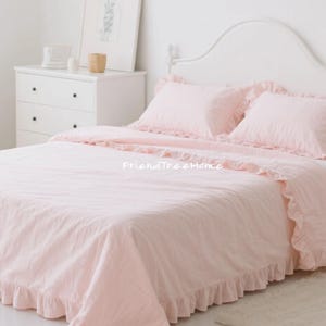 May include: A light pink bedspread and pillow set with ruffled edges. The bedding is on a white bed frame with a matching headboard. The room has a white dresser and a framed picture on the wall. The overall theme is a cozy bedroom.