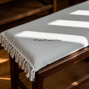 May include: A light gray linen bench cushion with a white fringe border. The cushion is on a dark wood bench. The text "FriendTreeHome" is printed on the cushion.