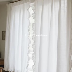 Op de afbeelding: Witte, doorschijnende gordijnen met een gerimpelde rand. De gordijnen hangen in een raam en laten natuurlijk licht door.