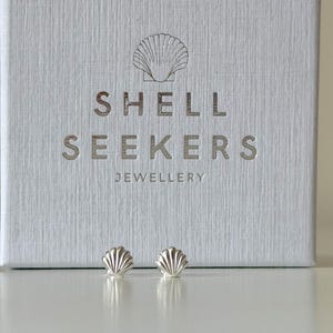 May include: Silver seashell stud earrings presented in front of a white box. The box is embossed with the words "SHELL SEEKERS JEWELLERY" and a seashell design. The earrings are small, detailed, and reflect light.