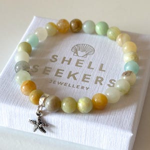 May include: A beaded bracelet with a starfish charm. The bracelet features a variety of earth-toned beads in shades of yellow, green, and brown. The charm is a small, silver-colored starfish. The bracelet rests on a white box with the text "SHELL SEEKERS JEWELLERY".