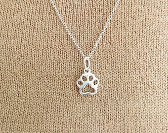 May include: A silver pendant necklace featuring a paw print design. The pendant is suspended from a delicate chain. The necklace is displayed against a tan sweater.