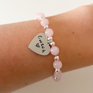 Könnte beinhalten: Ein rosa Perlenarmband mit einem silbernen Herzanhänger, auf dem "cute" steht.