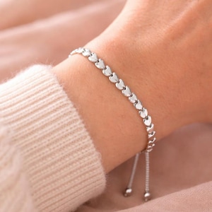 May include: A silver bracelet with heart-shaped links. The bracelet has an adjustable sliding bead closure. The jewellery is shown on a wrist, set against a soft pink backdrop.