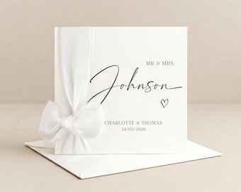 May include: A white wedding card with a ribbon and bow, featuring the names "Mr & Mrs Johnson" in elegant script, a heart symbol, and the date 24/05/2026. The card rests on a white envelope.