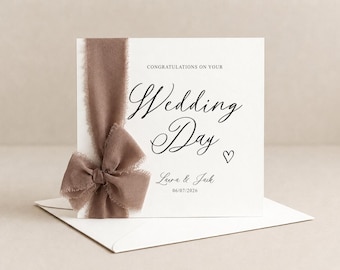May include: A square white wedding greeting card featuring the text "CONGRATULATIONS ON YOUR Wedding Day" with a small heart icon, the names "Laura & Jack," and the date "06/07/2026." A taupe-colored frayed silk ribbon is tied in a bow on the left side.