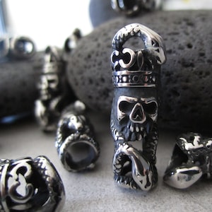 Peut inclure: Un charme en argent en forme de crâne avec un serpent enroulé autour, portant une couronne. Le charme est en métal et a un design détaillé.