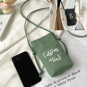 Puede incluir: Un bolso bandolera verde para teléfono con una correa larga. La bolsa tiene la inscripción "Custom Text" en blanco. Un smartphone negro está al lado de la bolsa. También se ve el texto "Available in Green or Black Gold or White Print".