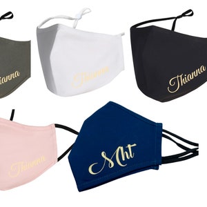 Könnte beinhalten: Fünf Stoffmasken mit verstellbaren Ohrschlaufen. Die Masken sind in verschiedenen Farben: grün, weiß, schwarz, rosa und blau. Jede Maske hat einen goldenen bestickten Namen. Die Namen sind "Thianna" und "Sht".