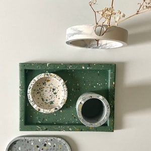 Puede incluir: Una bandeja verde con un tazón blanco y una taza blanca, ambos con un diseño de terrazo moteado. Hay una bandeja ovalada más pequeña con un diseño similar en primer plano.