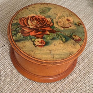 May include: A round wooden box with a smooth, light brown finish. The lid features a decorative print of a large, deep red rose and a smaller, pale yellow rose with green leaves against a muted, weathered-looking background with visible texture.