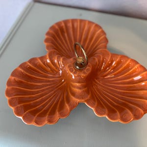 May include: An orange ceramic serving dish with three sections, shaped like stylized seashells. It has a gold-colored handle in the center. The dish is on a light blue surface.