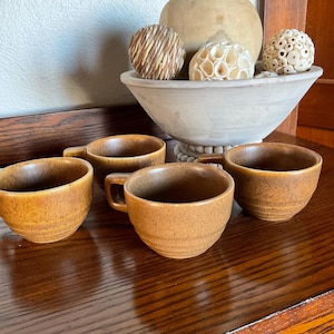 May include: Four brown ceramic coffee cups with handles are arranged on a wooden surface. The cups have a speckled texture and a rounded shape. A decorative bowl filled with decorative spheres sits in the background.