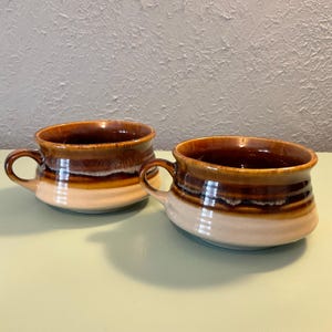 May include: Two ceramic soup bowls with handles. The bowls have a brown and tan glaze, with a darker brown interior. The bowls are round and have a classic design, suitable for serving soup or other dishes.