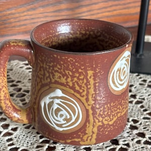 May include: A ceramic mug with a textured, reddish-brown and mustard-yellow mottled glaze. The exterior features two prominent circular motifs, each containing a hand-painted white spiral design. The mug has a sturdy handle and a cylindrical body.
