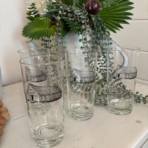 May include: Four clear glass tumblers with a black and white illustration of a barn on each. The glasses are on a white surface.