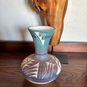 May include: A ceramic vase with a flared rim, featuring a gradient color scheme transitioning from teal at the top to dark purple and maroon at the base. The surface is decorated with white geometric line patterns and angular shapes.
