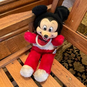 May include: A vintage Mickey Mouse plush doll. The doll has a black head with large ears, a white face, and a red body. It wears a bib with the word "Mickey" on it. The doll is sitting on a wooden surface.