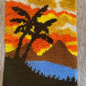 May include: A colorful rug with a silhouette of a palm tree in front of a sunset. The rug is made of a fluffy, textured material and features shades of orange, yellow, brown, and blue.
