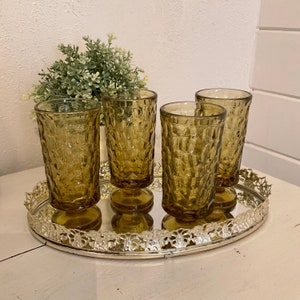 May include: Four amber glass tumblers with a textured design sit on a silver tray with a decorative edge. The tumblers have a footed base.