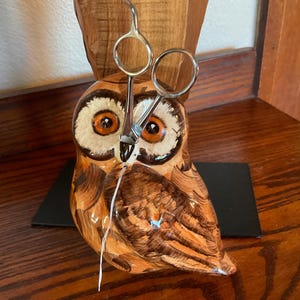 May include: A ceramic owl figurine with brown and orange hues, holding a pair of silver scissors. The owl has large, detailed eyes and is perched on a black base. The background features a wooden surface.