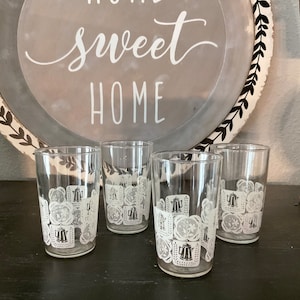 Op de afbeelding: Set van vier heldere glazen bekers met een wit ontwerp met een herhalend patroon van een theepot en andere keukengerei. De bekers staan op een donker houten oppervlak.