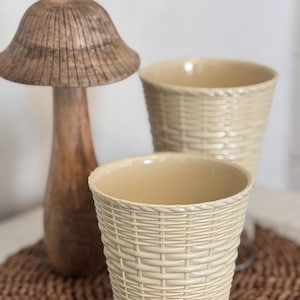 May include: Two cream-colored ceramic planters with a woven basket design sit on a brown woven mat. A wooden mushroom-shaped decorative piece stands to the left. The planters have a smooth interior and a textured exterior.