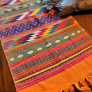 May include: A rectangular table runner featuring a vibrant woven pattern with horizontal bands of orange, yellow, blue, green, and magenta. The textile displays intricate geometric zigzags, diamond shapes, and small floral motifs with fringe ends.