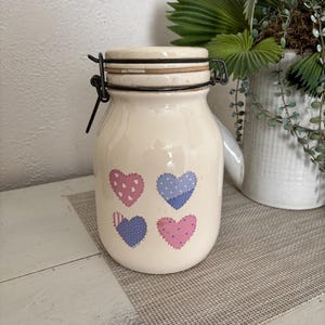 May include: A cream-colored ceramic canister with a wire-bail lid closure. The front of the jar features four heart-shaped designs in pink and blue, each decorated with small patterns like dots, crosses, and stripes, resembling fabric patches.