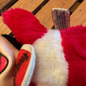 May include: Close-up of a plush toy featuring a red ear and a white and red foot. The toy has a tag that reads "KEY HOUSE PRODUCTIONS". The toy is made of soft, fuzzy material.