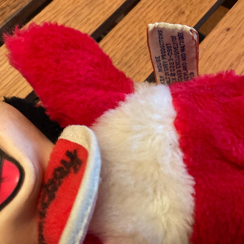 May include: Close-up of a plush toy featuring a red ear and a white and red foot. The toy has a tag that reads "KEY HOUSE PRODUCTIONS". The toy is made of soft, fuzzy material.