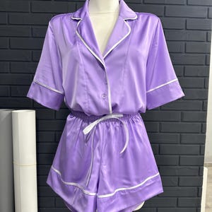 Puede incluir: Un conjunto de pijama de satén lila exhibido en un maniquí. El conjunto incluye una camisa abotonada de manga corta y pantalones cortos a juego. Ribetes blancos adornan los bordes de la camisa y los pantalones cortos. Un cordón blanco es visible en los pantalones cortos.