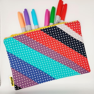 May include: A vibrant pencil case featuring a diagonal striped design in shades of red, blue, and purple, each adorned with white polka dots. The case is open, displaying several coloured markers. A yellow zip and a small label are visible.