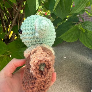 Puede incluir: Mu&ntilde;eca de ganchillo hecha a mano con forma de personaje con orejas grandes. La mu&ntilde;eca tiene una cabeza y orejas de color verde claro, un cuerpo marr&oacute;n y un ojo negro. La mu&ntilde;eca se sostiene en una mano sobre un fondo de hojas verdes.
