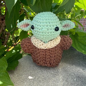 Puede incluir: Mu&ntilde;eco de ganchillo que se asemeja a un personaje de una popular serie de ciencia ficci&oacute;n. El mu&ntilde;eco tiene una cabeza verde claro con grandes ojos negros y orejas puntiagudas con detalles rosas. Viste un atuendo marr&oacute;n con cuello beige. Fondo de hojas verdes.