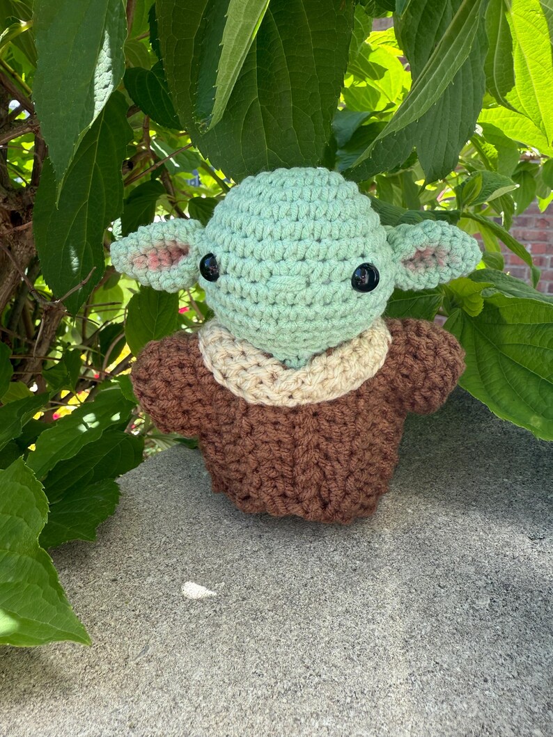 Puede incluir: Mu&ntilde;eco de ganchillo que se asemeja a un personaje de una popular serie de ciencia ficci&oacute;n. El mu&ntilde;eco tiene una cabeza verde claro con grandes ojos negros y orejas puntiagudas con detalles rosas. Viste un atuendo marr&oacute;n con cuello beige. Fondo de hojas verdes.
