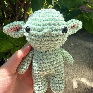 Puede incluir: Mu&ntilde;eco de ganchillo que se asemeja a un personaje popular, con cuerpo verde claro, ojos negros grandes y detalles rosas en las orejas. El mu&ntilde;eco hecho a mano mide aproximadamente 10 cm de altura, mostrando un patr&oacute;n de puntada detallado y texturizado.