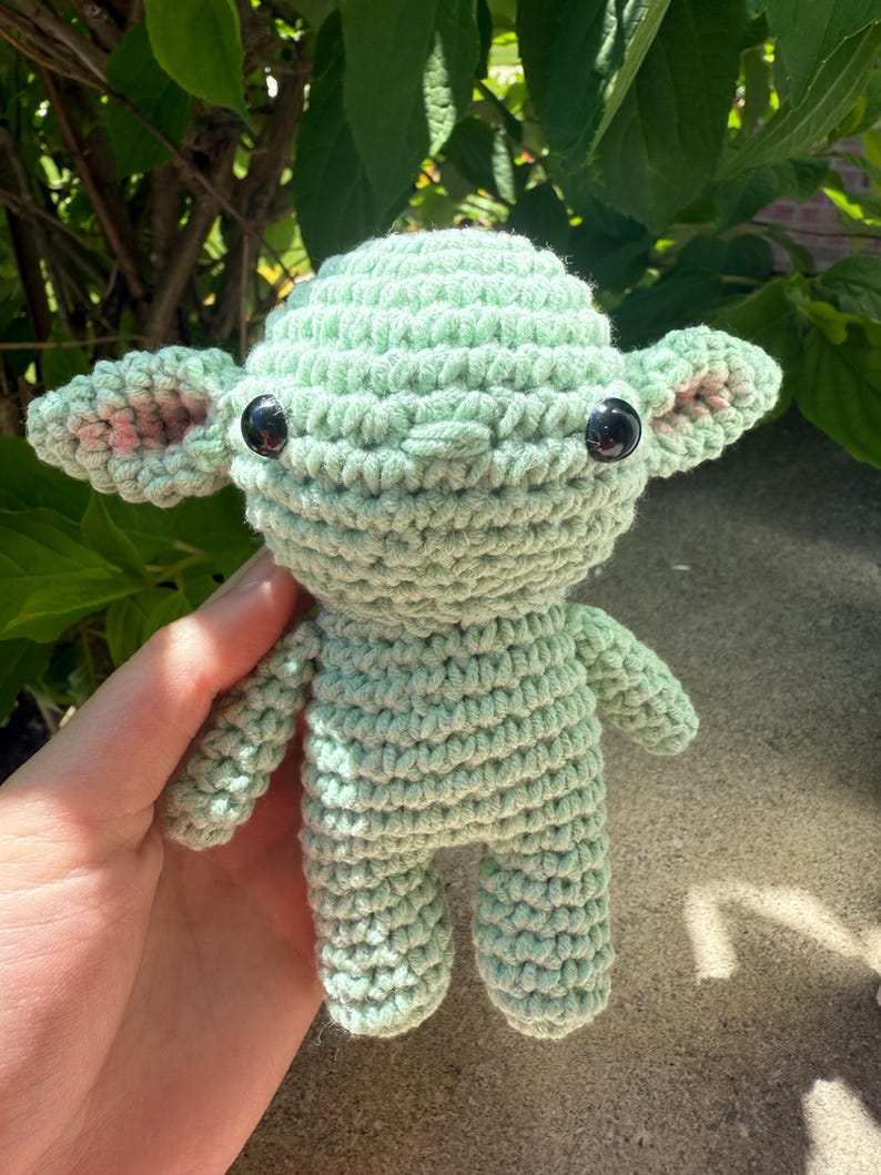 Puede incluir: Mu&ntilde;eco de ganchillo que se asemeja a un personaje popular, con cuerpo verde claro, ojos negros grandes y detalles rosas en las orejas. El mu&ntilde;eco hecho a mano mide aproximadamente 10 cm de altura, mostrando un patr&oacute;n de puntada detallado y texturizado.