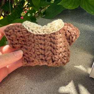 Puede incluir: Un peque&ntilde;o su&eacute;ter marr&oacute;n tejido a mano con un cuello color crema. La prenda en miniatura se sostiene en una mano, mostrando el detallado trabajo de puntadas. El su&eacute;ter est&aacute; dise&ntilde;ado para una mu&ntilde;eca peque&ntilde;a o un juguete.