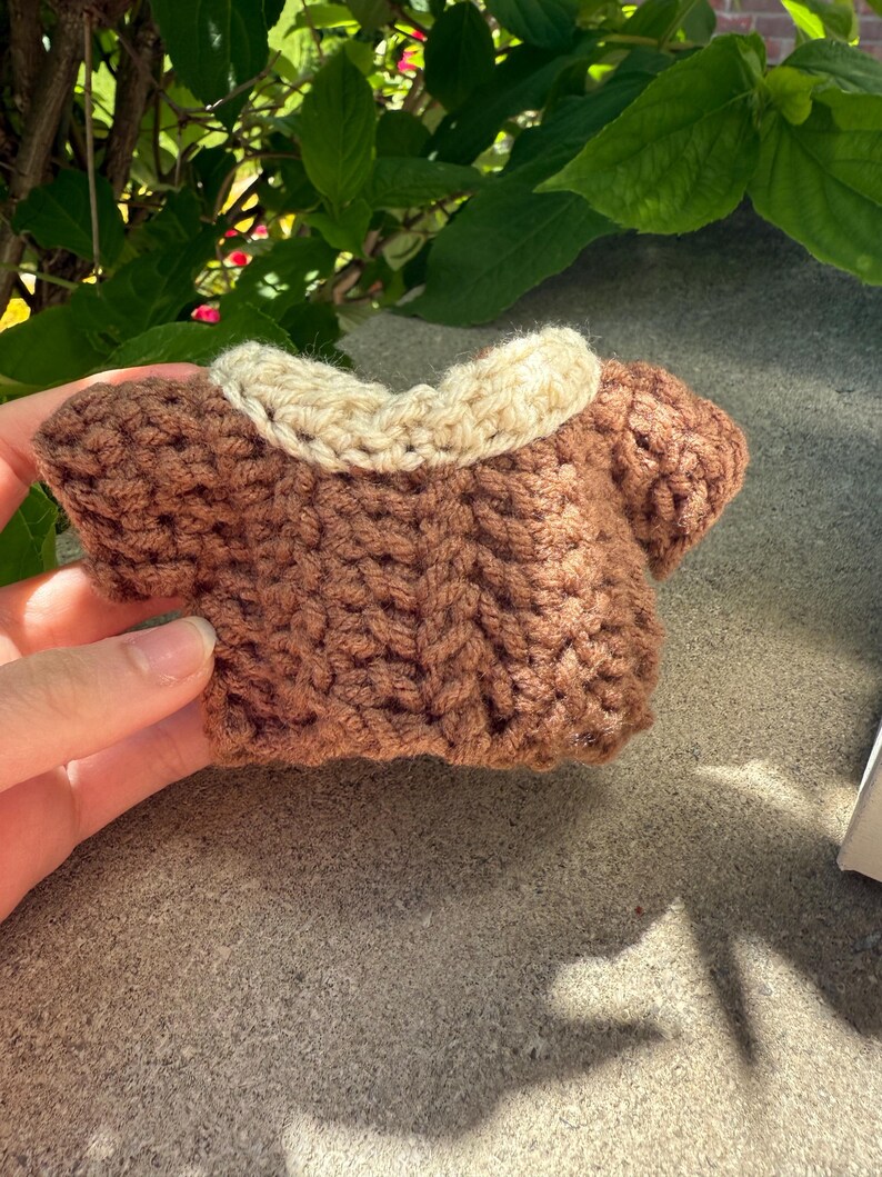 Puede incluir: Un peque&ntilde;o su&eacute;ter marr&oacute;n tejido a mano con un cuello color crema. La prenda en miniatura se sostiene en una mano, mostrando el detallado trabajo de puntadas. El su&eacute;ter est&aacute; dise&ntilde;ado para una mu&ntilde;eca peque&ntilde;a o un juguete.