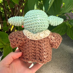 Puede incluir: Una mu&ntilde;eca de ganchillo de un personaje con orejas grandes y un abrigo marr&oacute;n. La cabeza de la mu&ntilde;eca es de color verde claro y el abrigo es marr&oacute;n con un cuello color crema. La mu&ntilde;eca se sostiene en la mano de una persona sobre un fondo de hojas verdes.