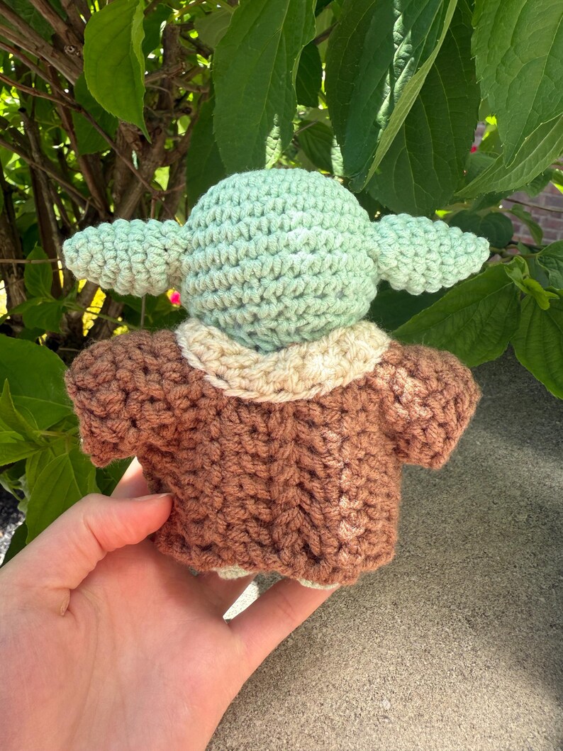 Puede incluir: Una mu&ntilde;eca de ganchillo de un personaje con orejas grandes y un abrigo marr&oacute;n. La cabeza de la mu&ntilde;eca es de color verde claro y el abrigo es marr&oacute;n con un cuello color crema. La mu&ntilde;eca se sostiene en la mano de una persona sobre un fondo de hojas verdes.