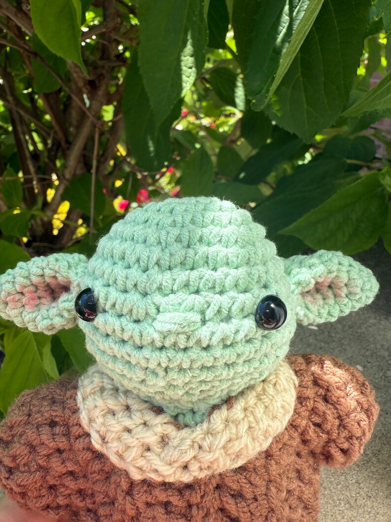 Puede incluir: Mu&ntilde;eco de ganchillo hecho a mano con una cabeza verde claro, ojos negros grandes y orejas grandes con detalles en rosa. El mu&ntilde;eco tiene un cuerpo marr&oacute;n con un cuello color crema. El fondo presenta hojas verdes y luz solar.