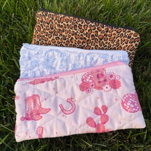 May include: Three rectangular quilted zipper pouches stacked together. The top pouch features a brown and black leopard print. The middle pouch has a light blue floral pattern. The bottom pouch is light pink with a western-themed graphic print.