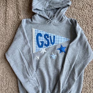May include: A gray hooded sweatshirt with a front pocket and drawstring. A blue and white pennant with the letters "GSV" is sewn on the front, along with blue and white stars. The sweatshirt is made of a soft material.