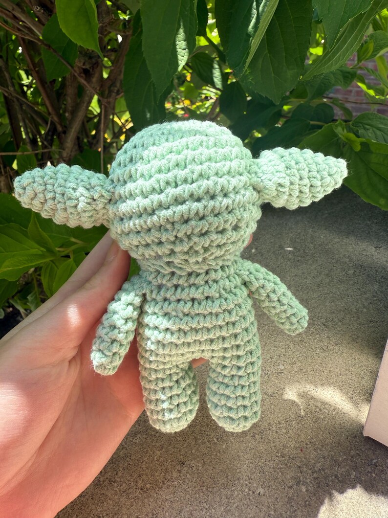 Puede incluir: Mu&ntilde;eco de ganchillo verde claro con orejas grandes y cuerpo peque&ntilde;o. El mu&ntilde;eco est&aacute; hecho a mano con una puntada texturizada, y los detalles incluyen brazos y piernas peque&ntilde;os. El mu&ntilde;eco es un personaje de una popular franquicia de ciencia ficci&oacute;n.