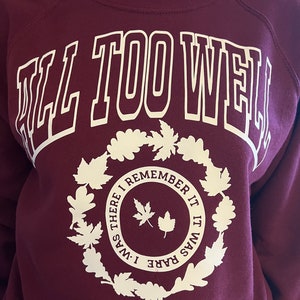 Könnte beinhalten: Burgunderfarbenes Sweatshirt mit dem Text "ALL TOO WELL" in großen weißen Blockbuchstaben. Darunter befindet sich ein weißer Blätterkranz, der einen Kreis mit dem Text "I WAS THERE I REMEMBER IT IT WAS RARE" umgibt.