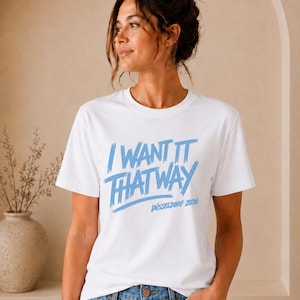Può includere: Una t-shirt bianca a maniche corte con il testo blu I WANT IT THAT WAY e DÜSSELDORF 2026 stampato sul davanti in un carattere stile pennellata. La maglietta ha un girocollo ed è abbinata a un paio di jeans blu.