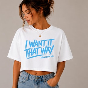 Könnte beinhalten: Ein weißes, kurzärmeliges bauchfreies Oberteil mit dem blauen Schriftzug I WANT IT THAT WAY und DÜSSELDORF 2026 auf der Vorderseite. Das T-Shirt hat einen Rundhalsausschnitt und eine lockere Passform, kombiniert mit einer blauen Jeans mit Metallknopf.