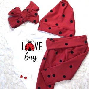 May include: A red and black ladybug-themed baby outfit set. The set includes a white onesie with "Love Bug" text and a ladybug graphic, a red bow headband, a knotted cap, and red pants with black polka dots.